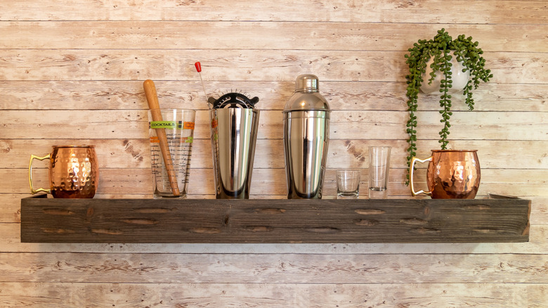 Dark wood wall mounted shelf holding cocktail shaker, shot glasses, tall glass with muddler, and mule mugs