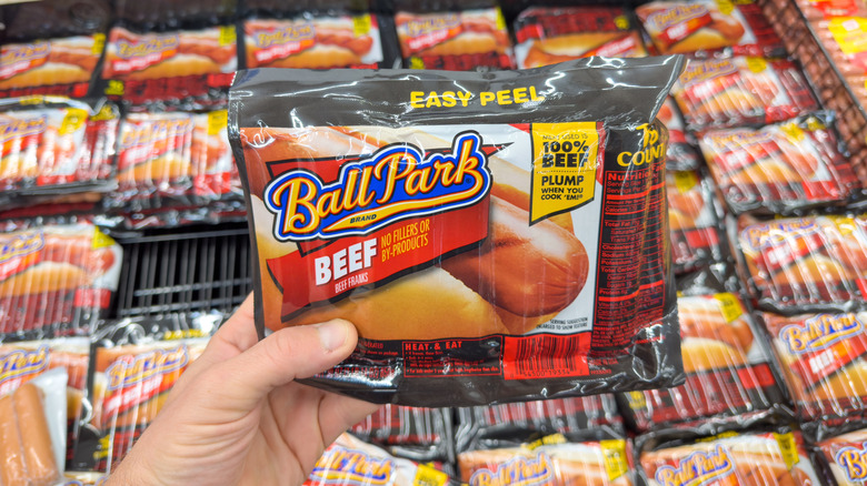 a hand holding up a package of ball park hot dogs above many other packages