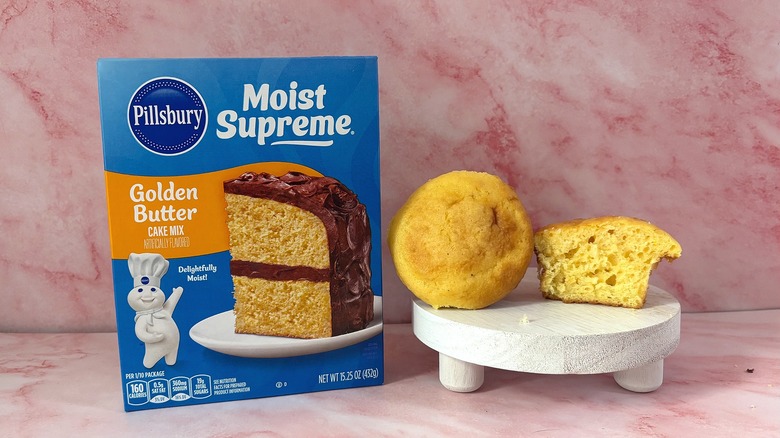 Pillsbury Golden Butter cake mix box next to two cupcakes on white platform, all on pink surface