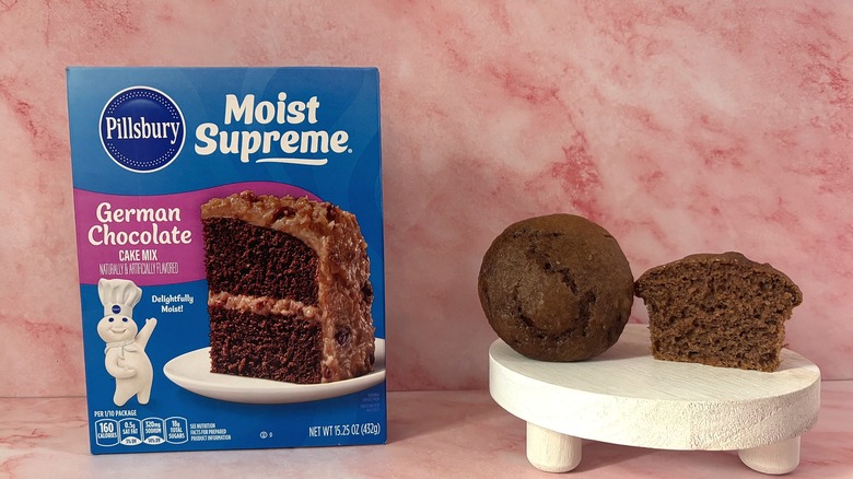 Pillsbury German Chocolate cake mix box next to two cupcakes on white platform, all on pink surface