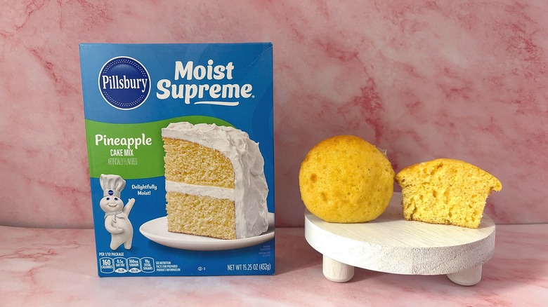 Pillsbury Pineapple cake mix box next to two cupcakes on white platform, all on pink surface