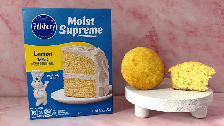 Pillsbury Lemon cake mix box next to two cupcakes on white platform, all on pink surface