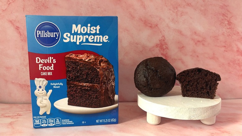 Pillsbury Devil's Food cake mix box next to two cupcakes on white platform, all on pink surface