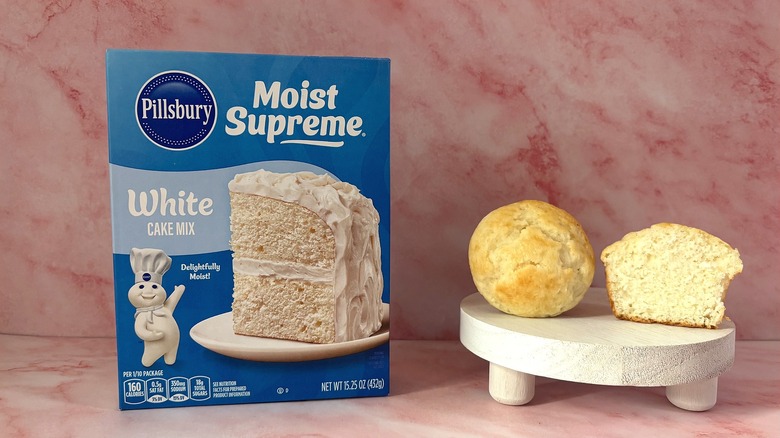 Pillsbury White cake mix box next to two cupcakes on white platform, all on pink surface