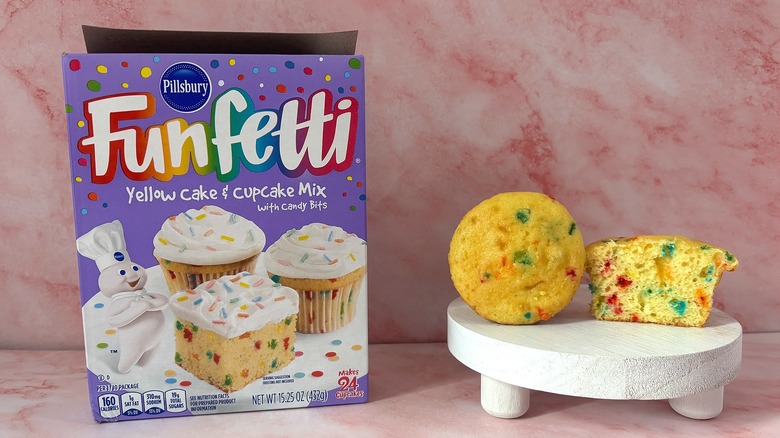 Pillsbury Funfetti cake mix box next to two cupcakes on white platform, all on pink surface