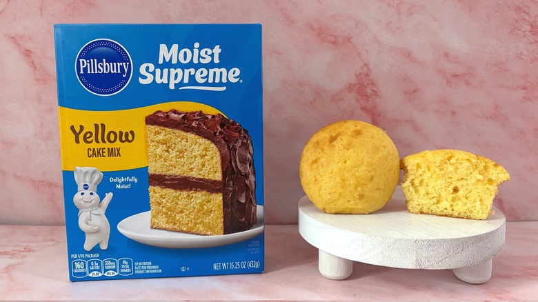 Pillsbury Yellow cake mix box next to two cupcakes on white platform, all on pink surface
