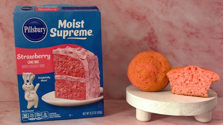 Pillsbury Strawberry cake mix box next to two cupcakes on white platform, all on pink surface