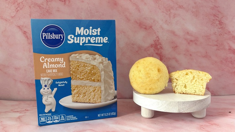 Pillsbury Creamy Almond cake mix box next to two cupcakes on white platform, all on pink surface