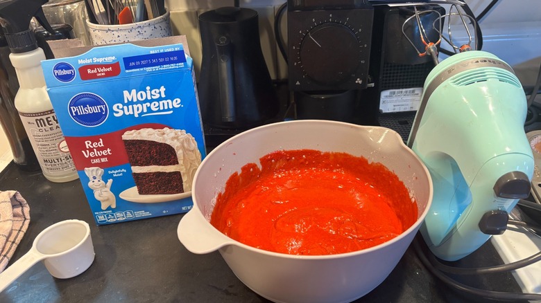 Counter with baking mix box, mixing bowl full of red batter, hand mixer, measuring cup