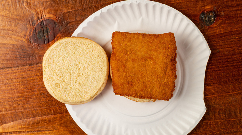 A breaded square fish fillet on a hamburger bun sitting on a paper plate