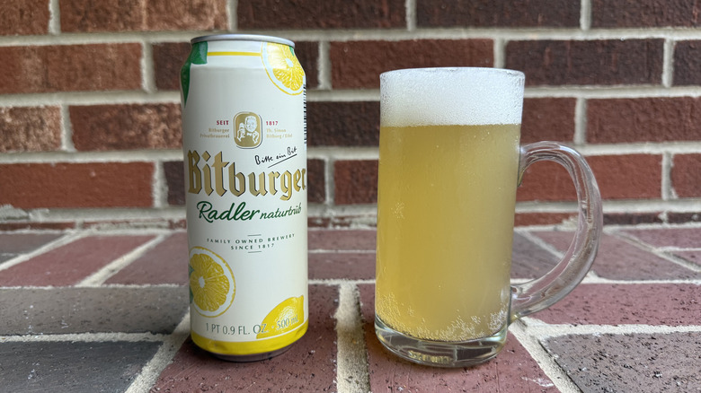Glass of pale yellow radler next to Bitburger can on brick surface