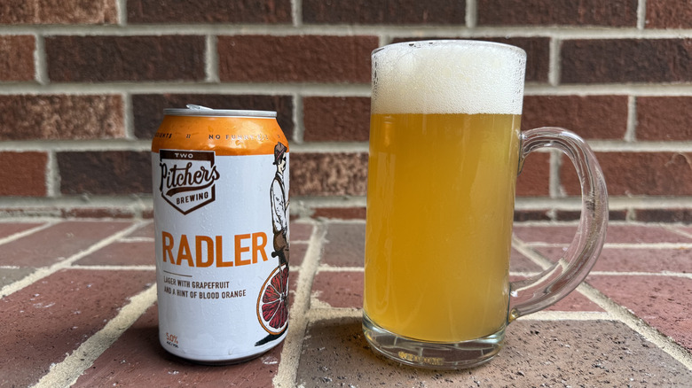 Glass of golden-hued radler next to Two Pitchers can on brick surface