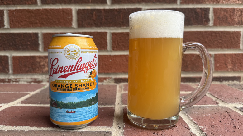 Glass of opaque golden-orange shandy next to Leinenkugel's orange shandy can on brick surface