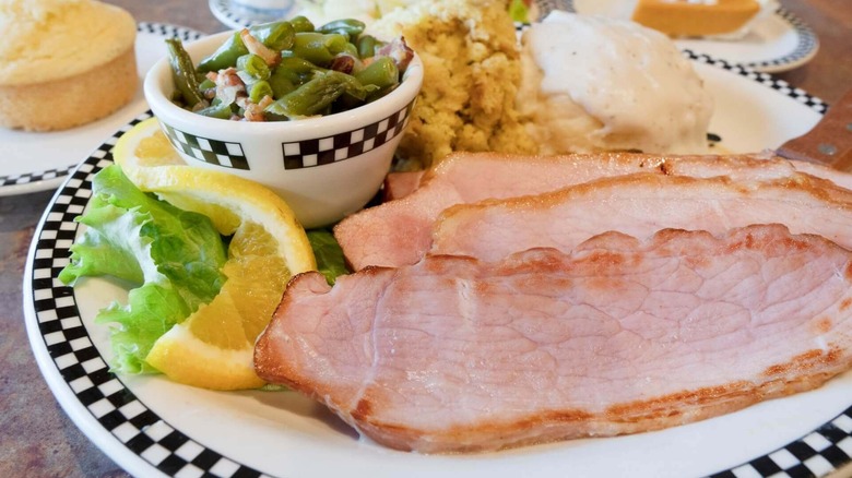 A plate of sliced ham with gravy, mashed potatoes, and green beans with a side salad and corn bread