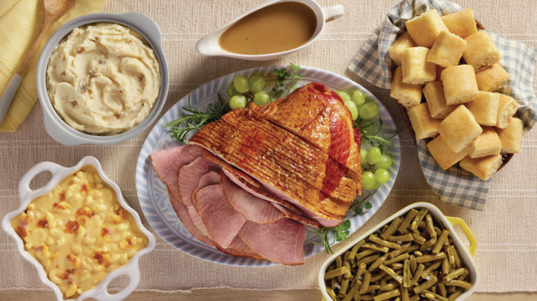 An Easter meal featuring sliced honey-glazed ham, mashed potatoes and gravy, green beans, mac and cheese, and fresh bread