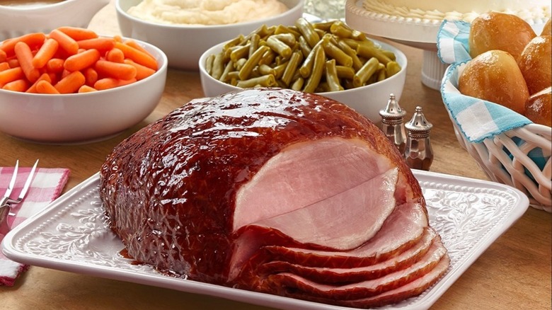 A dinner table set with sliced, glazed ham, green beans, rolls, carrots, and mashed potatoes
