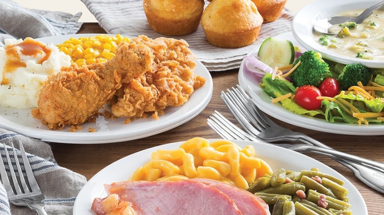 A restaurant table with plates of ham, fried chicken, green beans, veggies, soup, and corn bread