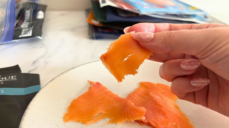 Hand holding piece of smoked salmon above plate of smoked salmon, smoked salmon packages in the background
