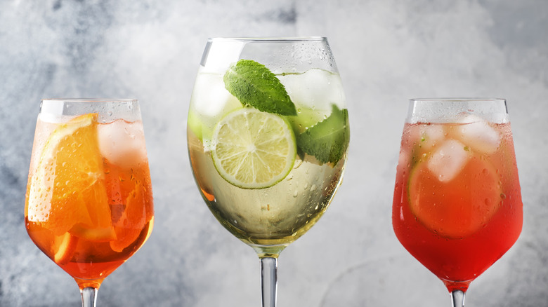 Three Italian Spritz cocktails in tall wine glasses with ice cubes and colorful fruit and herb garnishes