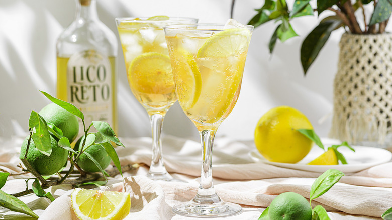 Two glasses of Limoncello Spritz with lemon slices with a bottle of limoncello in the back and lime and lemons lying around