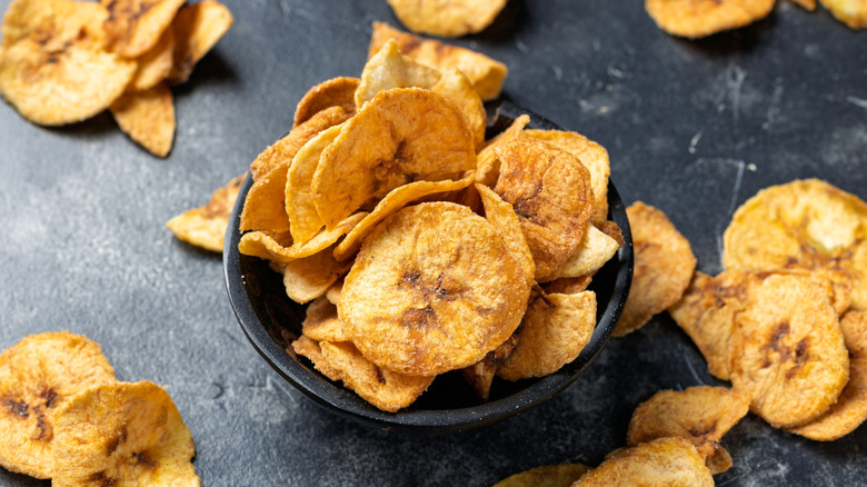 Banana chips in a black bowl and around it, on black surface