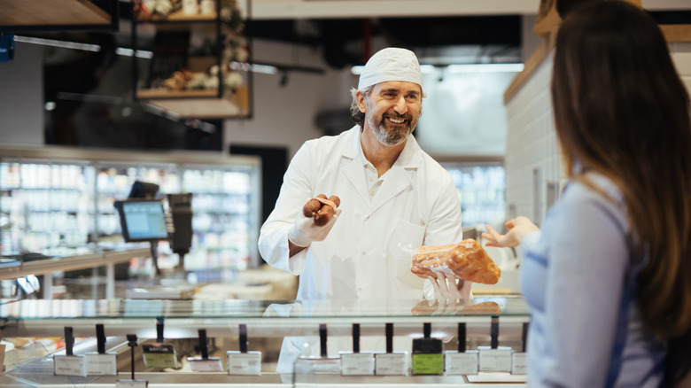 Butcher behind counter holding meat and talking to female customer.
