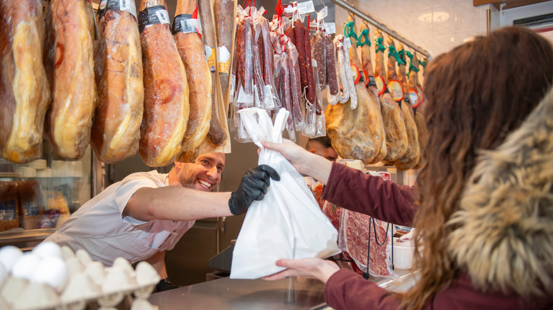 Butcher behind a counter with hanging cured meats handing a woman a bag