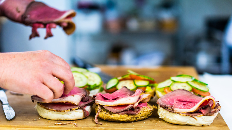 Hands preparing multiple fresh deli sandwiches on cutting board.
