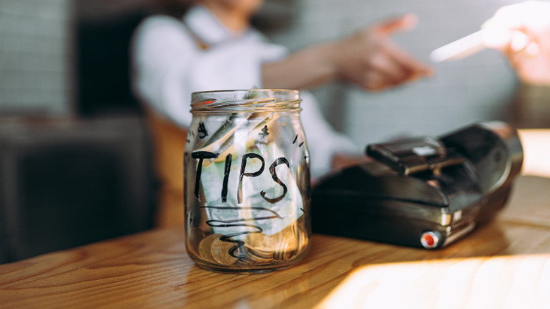 A glass jar that says "Tips" on it in black sharpie pen on a wooden counter