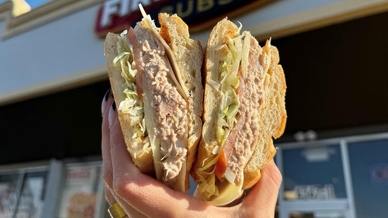 Two halves of a tuna sandwich being held up by a hand in front of a Firehouse Subs