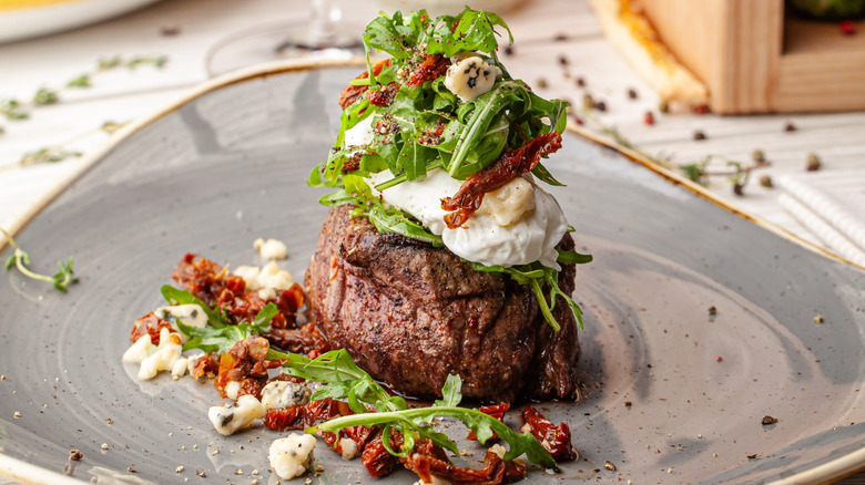 Filet steak topped with burrata, arugula, sundried tomatoes, and goat cheese on gray plate