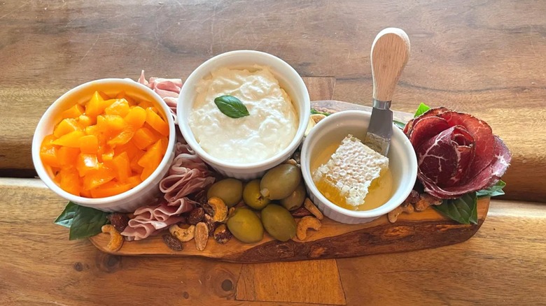 Wooden cheese board with ramekins of stracciatella cheese, honeycomb, olives, nuts, and cured meats