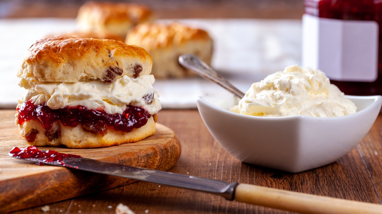 Scone spread with raspberry jam and creamy filling next to bowl of cream
