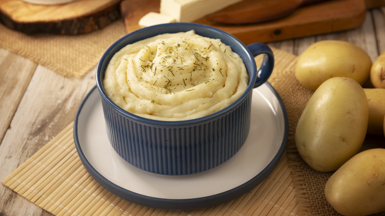 Blue ramekin full of mashed potatoes on saucer next to whole white potatoes