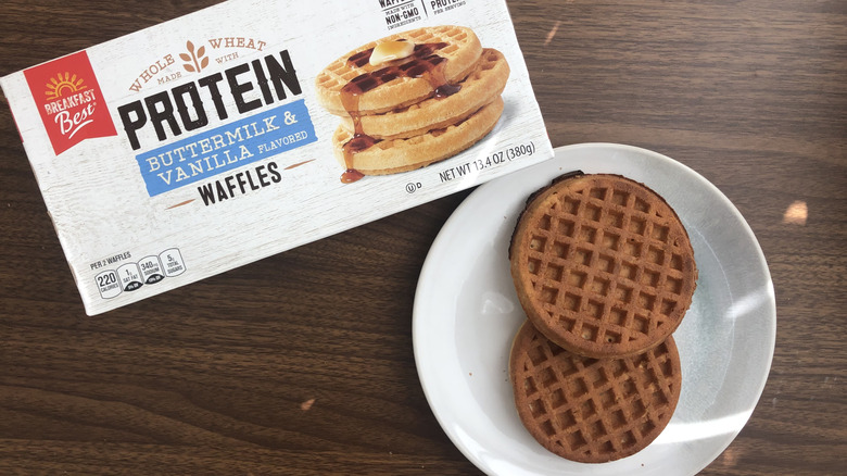 A box of Aldi Breakfast Best buttermilk and vanilla protein waffles next to a white plate with two waffles on it