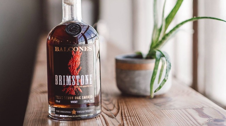 A bottle of Balcones Brimstone placed on a wooden shelf with a blurred plant shown in the background