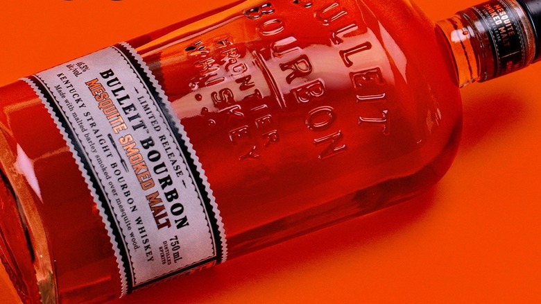 A close up of a bottle of Bulleit Mesquite Smoked Malt laid down on an orange background