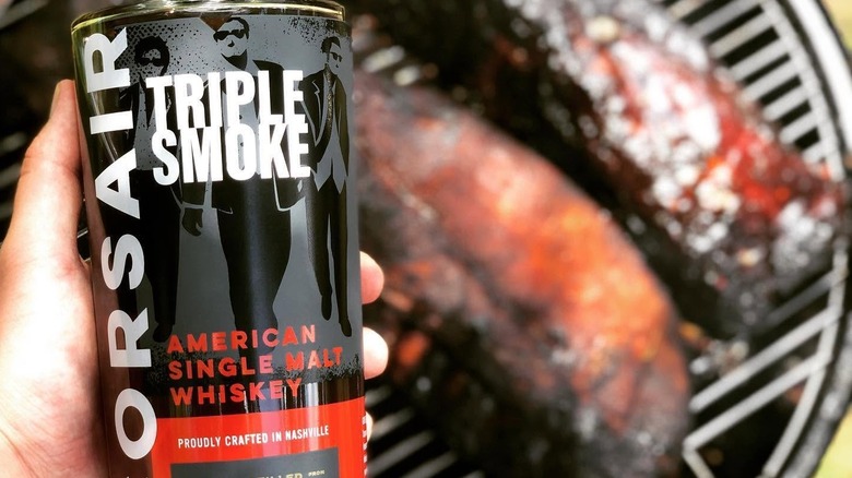 A bottle of Corsair Triple Smoke held up the the camera above meats cooking on a barbecue below