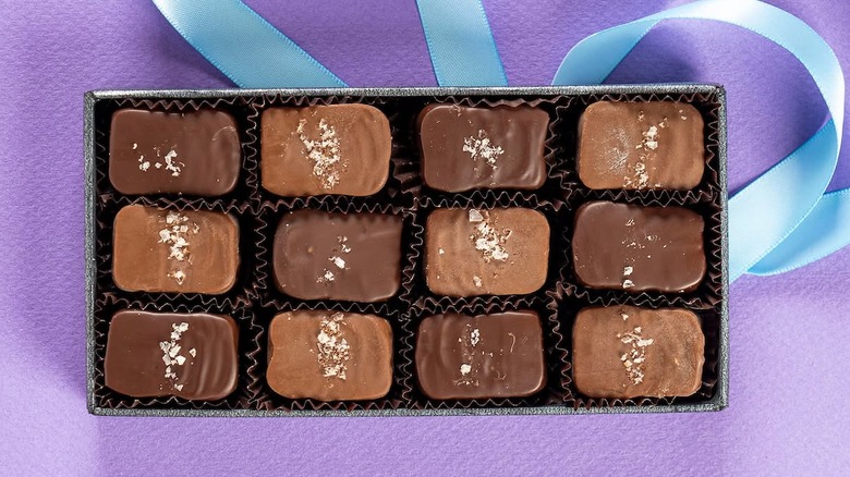 Open box of sea salt-topped chocolates on purple background atop pale blue ribbon