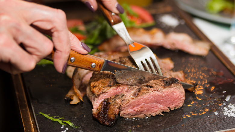 A person cutting through steak with a knife and fork on a cutting board