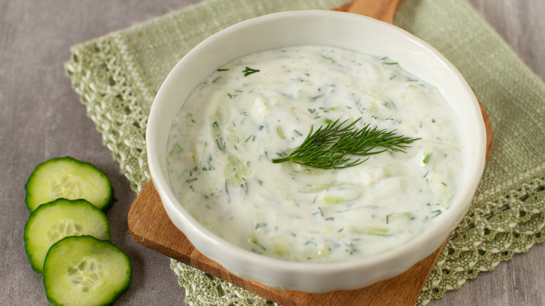 Bowl of tzatziki garnished with sprig of dill, next to slices of cucumber