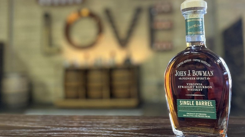 A bottle of John J. Bowman Single Barrel on a wooden surface in front of a blurred rustic background