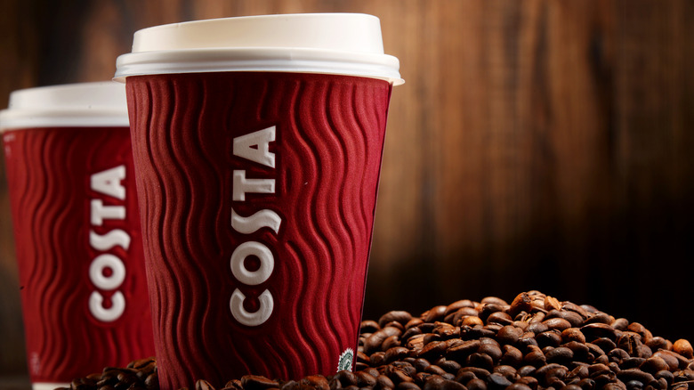Two Costa-branded disposable coffee cups next to whole coffee grounds.