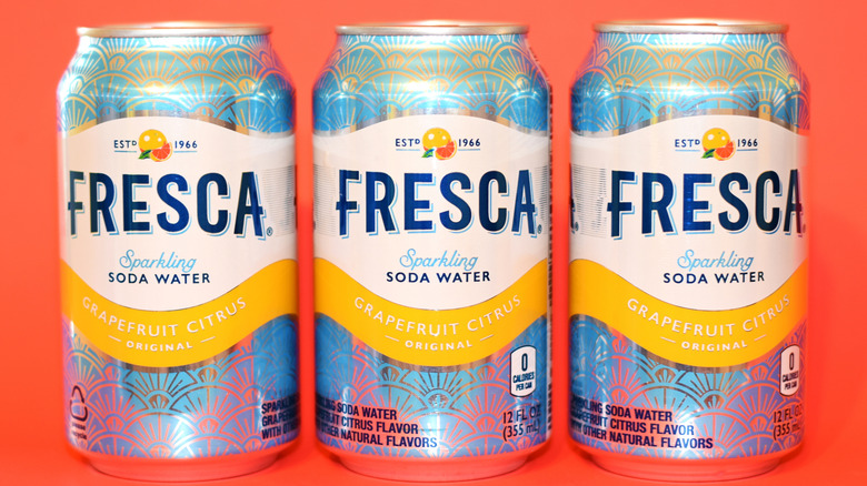 Three cans of Fresca sparkling soda water on display against an orange background.