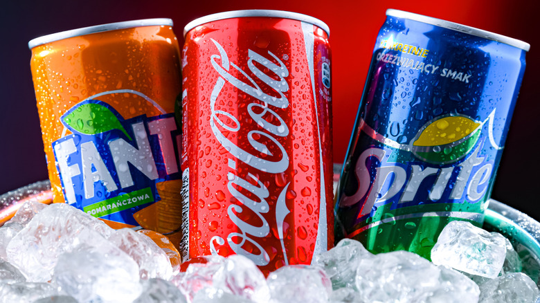 A can of Coca-Cola flanked by cans of Fanta and Sprite inside ice, with condensation.