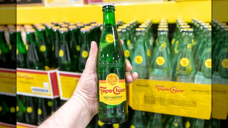 A hand holding a green glass bottle of Topo Chico, with other bottles blurred in the background.