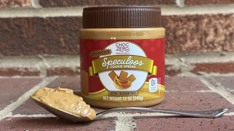 Jar of ChocZero crunchy speculoos cookie spread behind spoonful of cookie butter on brick surface