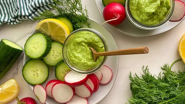 A bowl of green dip on a plate of cucumber slices, radishes and fresh dill.