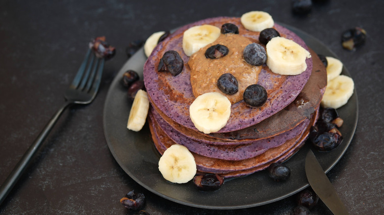 A plate of stacked ube pancakes topped with bananas and bluberries