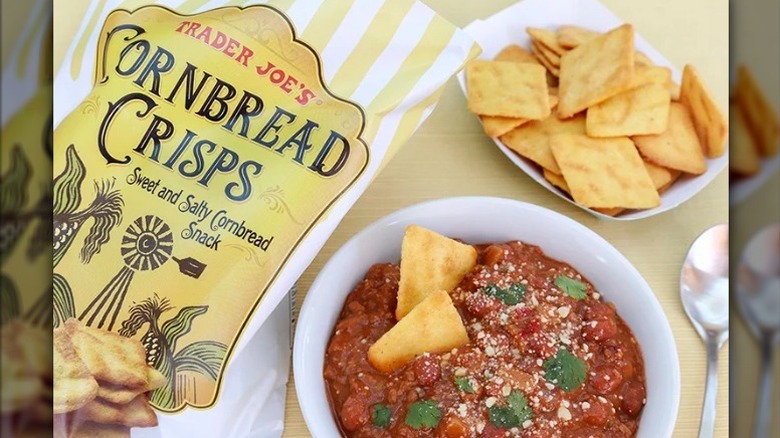 Trader Joe's cornbread crisps in bag and bowl with a bowl of chili and spoon on a pale yellow background.